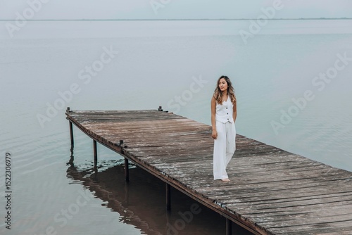 Asian woman in white outfit stands on wooden pier, calm lake, peaceful overcast, viewed from behind with contemplative mood, mindfulness, travel retreats, wellness