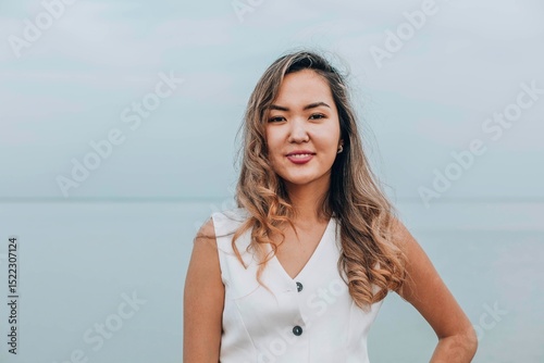 Asian woman smiling and looking at camera on a calm lakeside, dressed in white outfit, displaying a confident and cheerful emotion, concept of wellness, skincare, personal development