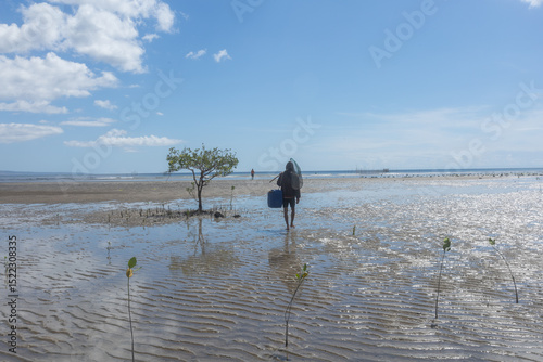 A fisherman walks on foot through the newly planted mangrove area, carrying makeshift fishing gear, hoping for a bountiful catch at his fish cage location