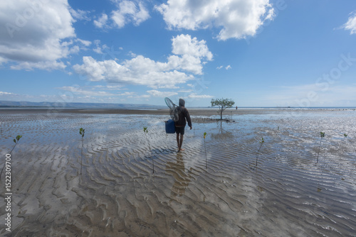 A fisherman walks on foot through the newly planted mangrove area, carrying makeshift fishing gear, hoping for a bountiful catch at his fish cage location