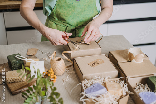 Person arranges kraft soap boxes on a table with shredded filler and handmade product tags. Handmade movement, analog creativity, DIY packaging, human touch in business