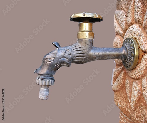 A garden tap shaped like an animal head, mounted on a reddish-white terracotta wellhead, with a partial handle, close-up shot, against a gray, monochrome background.
