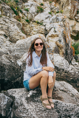 Young happy brunette woman in sunglasses, blue shirt, denim shorts and sandals sitting on rocks and smiling