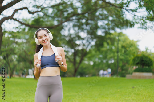 Happy asian sportswoman wearing headphones and holding towel, enjoying break after exercising outdoors in a park with green grass and trees