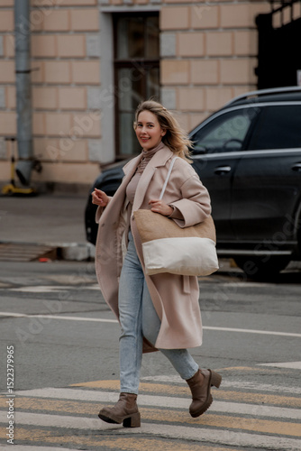 A young Caucasian woman with long blonde hair walks briskly across a street in Saint Petersburg. She wears a light coat and carries a large bag.