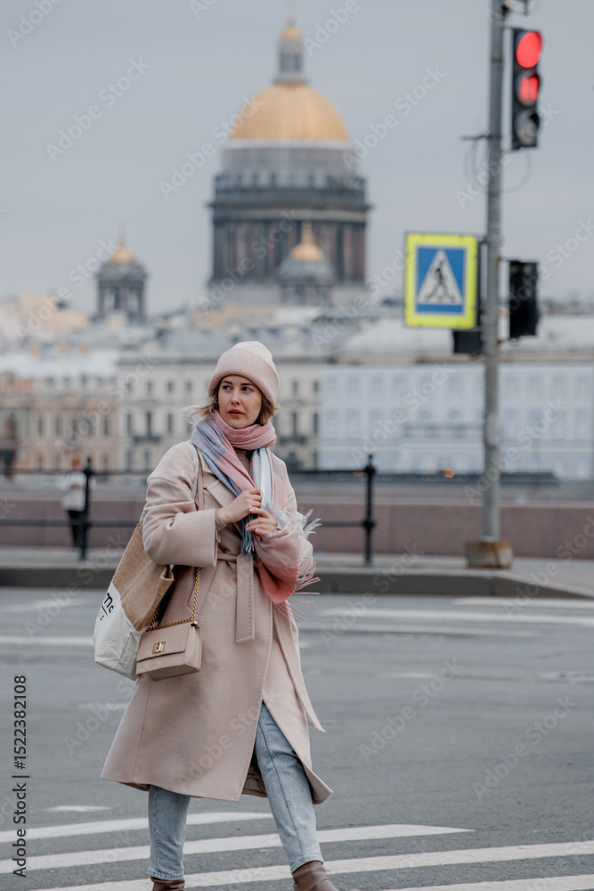 Fototapeta premium Saint-Petersburg, Russia, 06.06.2025.A young Caucasian woman with long blond hair walks across a street in Saint Petersburg. The golden dome of St. Isaac's Cathedral is visible in the background.