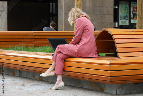 Blonde woman sitting with laptop on wooden bench on a street. Working in summer city