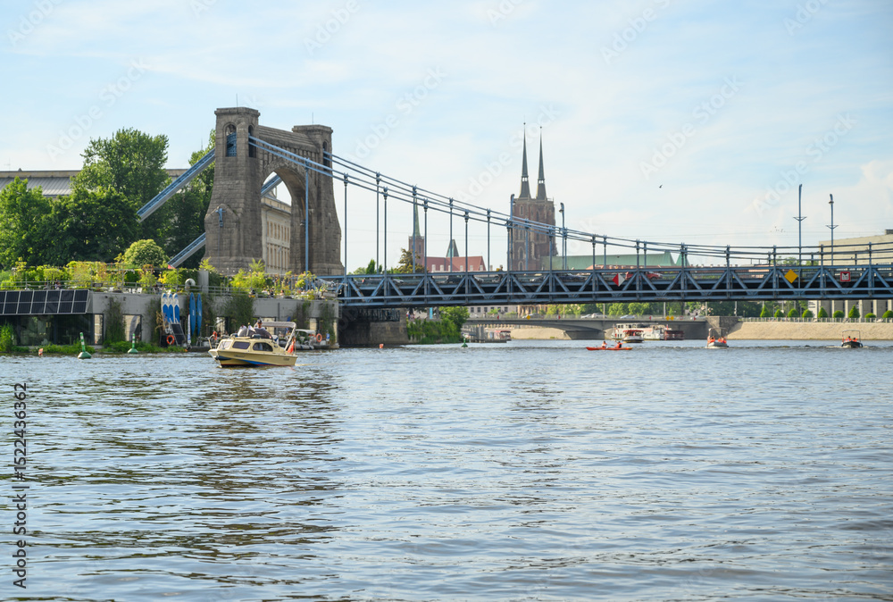 Obraz premium Urban river scenery featuring the Grunwaldzki suspension bridge and neo-Gothic cathedral on a clear afternoon in Wroclaw, Poland.