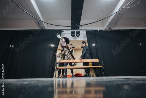 A group of young actors portrayed onstage preparing for a theatrical production, highlighted against a dramatic backdrop on a well-lit theatre setup showcasing their artistry and expressions.