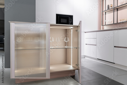 kitchen cabinet with glass doors and light inside. Beautiful wine glasses on shelves. close-up. stylish modern built-in kitchen with cabinets. kitchen facades are white and peach. kitchen unit