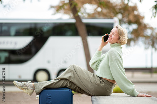 Smiling woman with short blond hair seated with suitcase while talking on phone outdoors