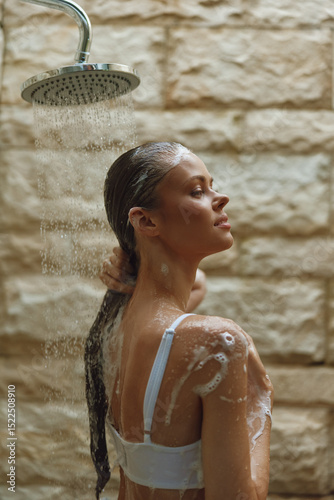 woman showering outdoors with soap suds on skin, wet hair, natural stone wall background, relaxed mood, warm lighting, personal care moment