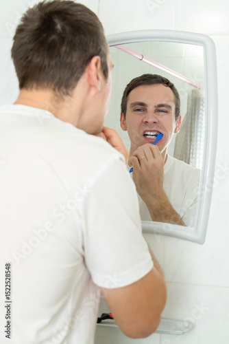 Young man brushing teeth with toothbrush while standing before mirror at home in the bathroom