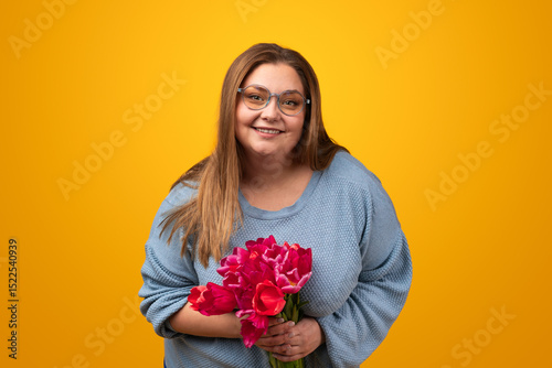 Optimistic adult obese female with long brown hair in gray sweater and eyeglasses, smiling and looking at camera while standing against yellow background with bunch of fresh tulips