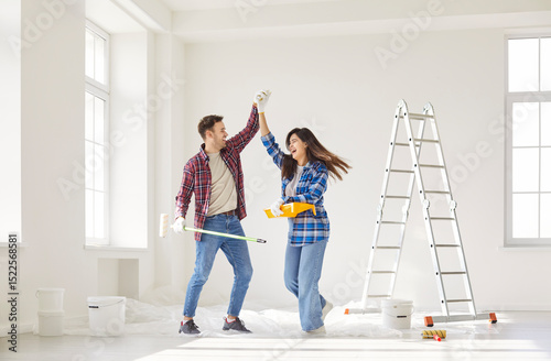 Happy family couple celebrating by dancing together while painting walls during the renovation of new home. Their joy reflects teamwork, construction efforts, and shared happiness.