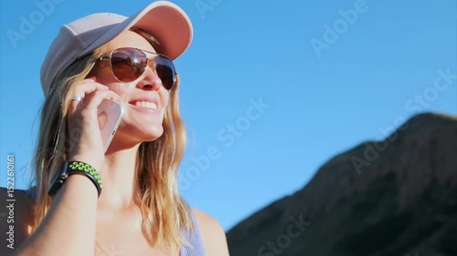 A beautiful, upbeat woman with loose hair, a pink cap, and sunglasses smiles while talking on the phone against a mountain backdrop. Sunlight lights her face, reflecting the joy of the conversation.