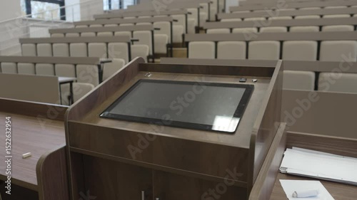 View from modern podium with digital tablet overlooks spacious lecture hall. Space filled with soft chairs and high-tech presentation setup