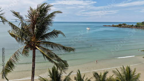 Fototapeta Naklejka Na Ścianę i Meble -  Lush palm trees framing a picturesque tropical beach with tourists swimming and sunbathing in crystal clear turquoise water and a yacht sailing in the distance