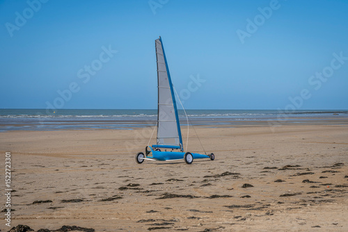 Asnelles-Sur-Mer, France - 05 16 2025:  View of a white and blue competition land yacht, a wheeled vehicle propelled by a sail along the beach between sand and sea