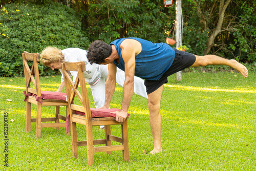 Senior couple doing balancing yoga pose in garden using chairs