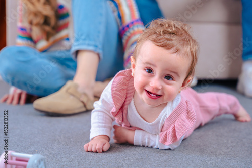 Adorable baby crawls on carpet smiling at the camera, showing joy and curiosity. Mother sits behind on the floor. Sweet indoor family moment filled with warmth and natural light. 