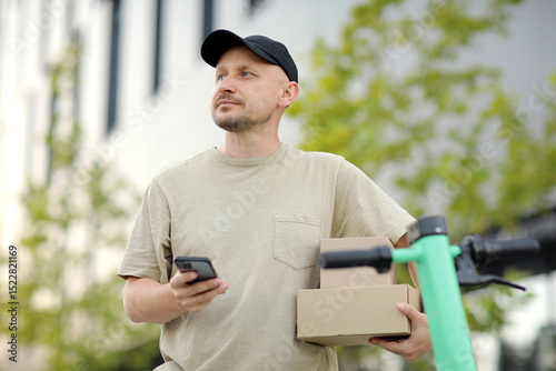 Portrait of young man using phone holding cardboard boxes near parked electric scooter on street at business area outdoors. Concept of sustainability, delivering and eco-friendly modern lifestyle