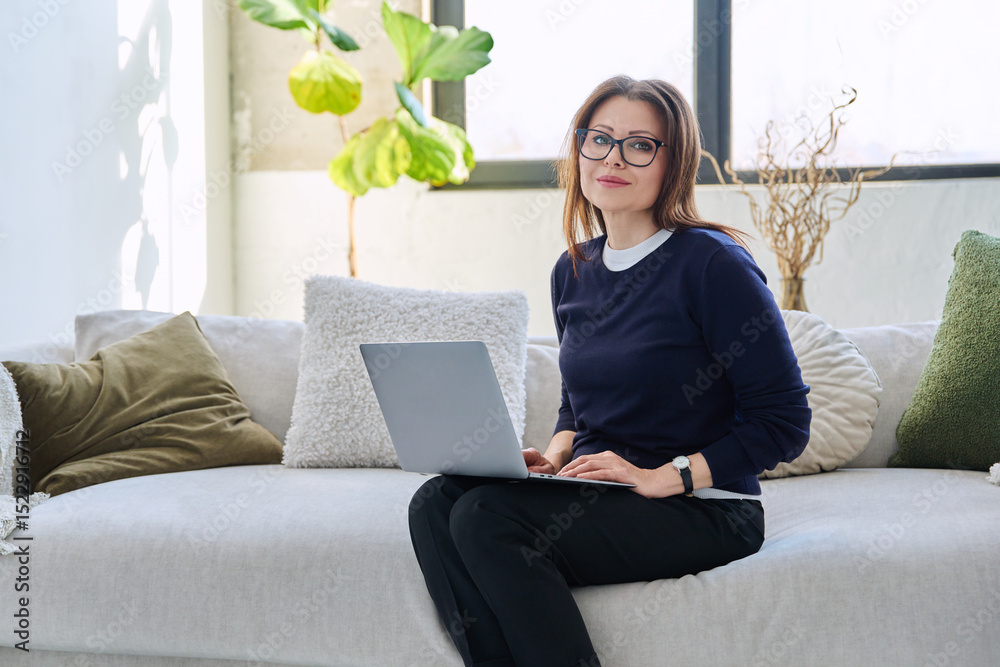 Naklejka premium Portrait of middle aged woman with laptop sitting on sofa at home