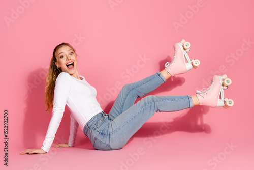 Fotografie Cheerful young woman in casual denim and pink roller skates sitting on a pink background with an excited and happy expression