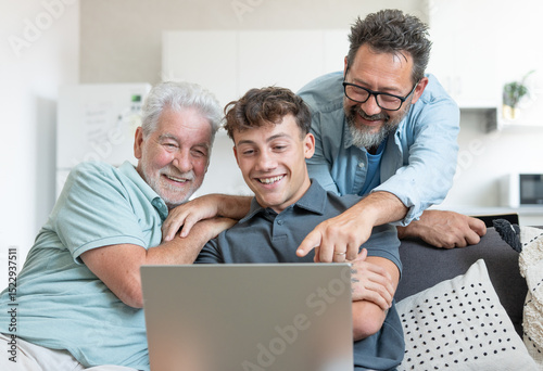 Happy multigenerational family sitting on sofa using laptop together - grandfather with mature son and young grandson enjoying carefree moments together learning use of computer