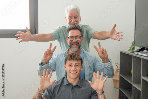 Happy multigenerational family of males stay together posing on scale for memory gesturing positive sign. Grandfather, mature son and young grandson laughing while looking at camera
