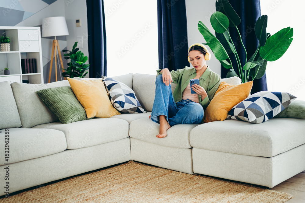 Obraz premium Charming woman relaxing on a stylish sofa with pillows and houseplants, enjoying a moment of leisure while using her smartphone