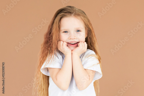 Headshot of pleased high spirit pretty female model, has warm pleasant smile, keeps hands under chin, wears white t-shirt, isolated over brown background. Happiness concept
