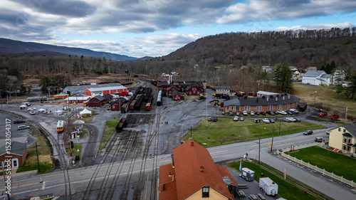 A view of a historic railway station surrounded by vintage train cars and buildings nestled in a valley. The location is peaceful, with mountains in the background under a partly cloudy sky.