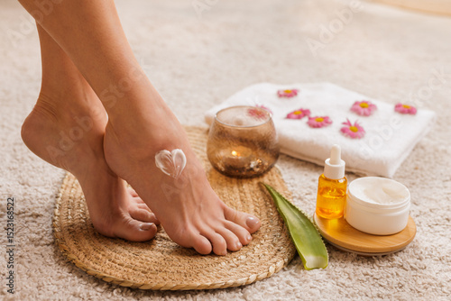 Woman’s feet with moisturizing cream in heart shape on skin, surrounded by aloe leaf, oil, and towel with flowers in a serene home spa setting, promoting self-care and natural beauty.