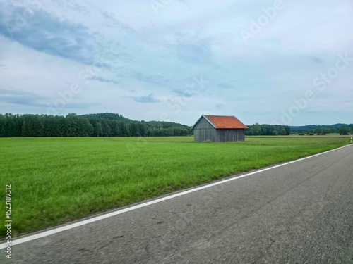 Quiet roadside view of a small wooden cabin amid green fields under a cloudy sky