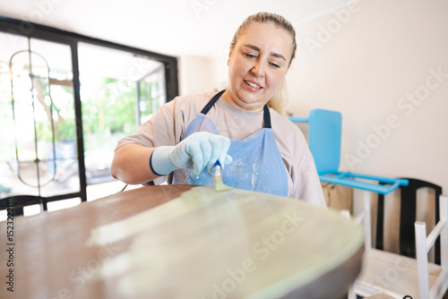 Woman in a small workshop restoring an old wooden chair, wearing a brown shirt and gloves, painting the chair. Small business in garage