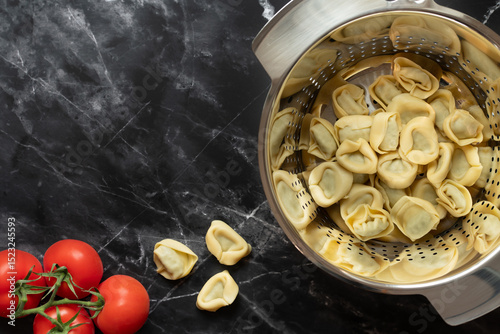 Freshly cooked tortellini are in a metal colander. The colander stands on a dark marble plate in the kitchen. There are still tomatoes on the plate. There is space for text.