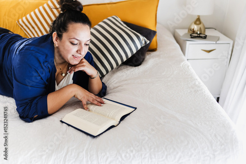 Young latin woman plus size reading book and lying on bed at bedroom in home in Mexico Latin America, hispanic overweight female	