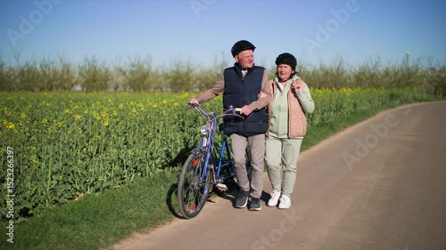 An elderly couple dismount from bicycle walks along a road in the countryside. 