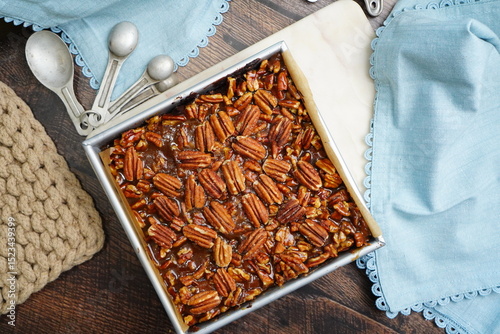 Decadent Pecan Brownies with caramel in a baking pan with light blue napkins.  Freshly baked brownies in a pan with measuring spoons and napkins on a wood counter top.