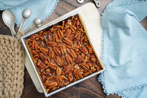 Decadent Pecan Brownies with caramel in a baking pan with light blue napkins.  Freshly baked brownies in a pan with measuring spoons and napkins on a wood counter top.