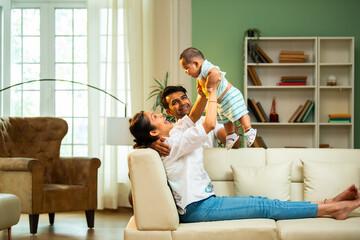 Parents catch baby boy while sitting on sofa at home, enjoying fun playful family bonding time