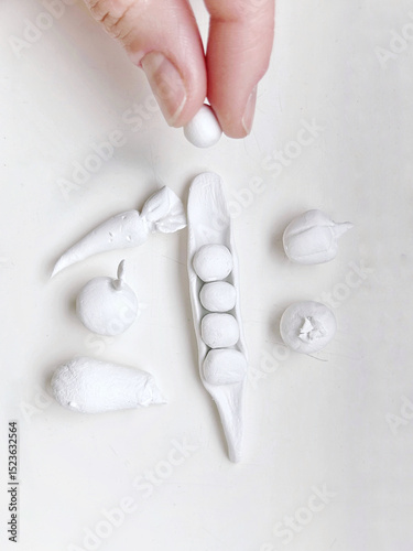 A hand arranges tiny handcrafted clay fruits and vegetables on a white surface
