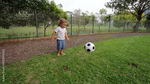 Barefoot toddler drops soccer ball on grass and kicks it forward with surprising ease and control. Captured in slow motion during evening game at fenced dog walk area