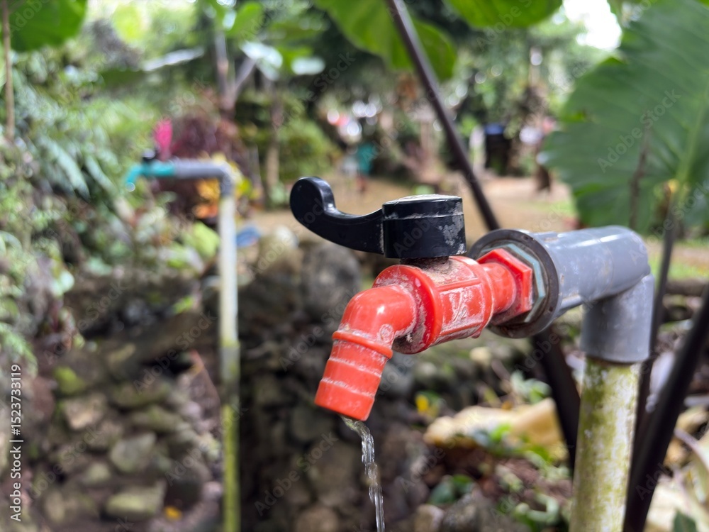 Fototapeta premium A close-up shot of a red plastic outdoor faucet with a black handle, set against a background of lush green plants and natural stone