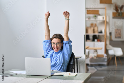 Excited office worker winner using laptop computer celebrating goal achievement. Happy young woman student or employee winning online getting good news in email raising hands feeling euphoric. photo
