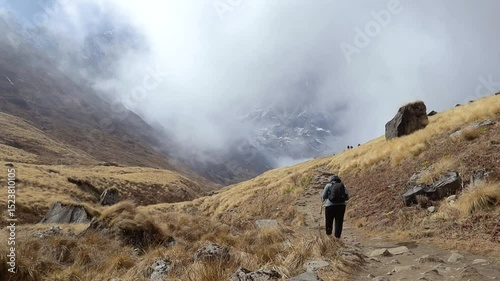 Solitary female trekker walking through a mist-covered trail  of Annapurna base camp trek in Nepal, surrounded by dry golden grass and rugged terrain in the Himalayas.
