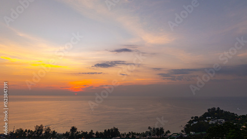 A breathtaking aerial view of Kata Beach in Phuket, Thailand, captured during sunset. The image showcases the coastline, small islands, lush green hills, and the vibrant cityscape under a bright sky