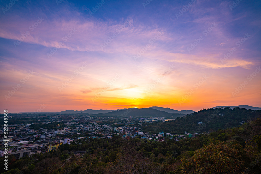 Fototapeta premium A scenic view of a town nestled among lush green mountains at sunset, with a temple structure perched on the hillside and a vibrant orange sky creating a warm, tranquil atmosphere.