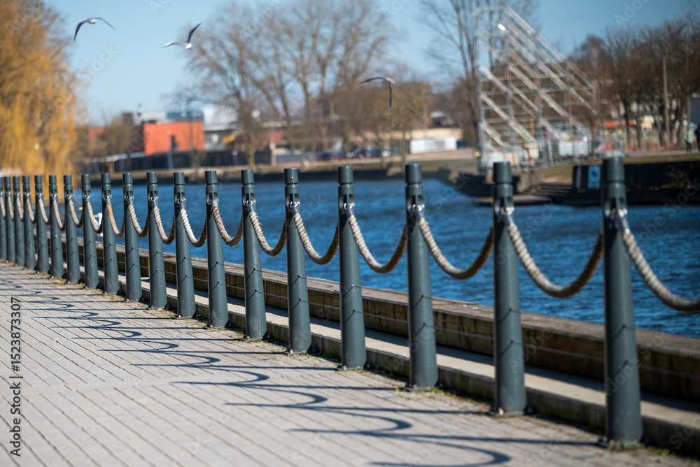 Naklejka premium A riverside promenade features a row of black metal posts connected by thick ropes. The walkway is paved with wooden planks. Birds are flying above, and the river runs alongside, with several bare tre